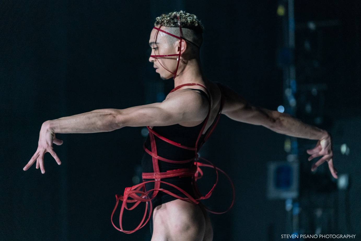 A dancer in a black leotard and red ribbon outstretches their arm their wrists bent.